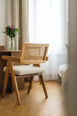 Ein Holzhocker mit gewebtem Rücken und gepolstertem Sitz, platziert in einem Raum mit Holzboden und einem Fenster mit durchsichtigen Vorhängen.