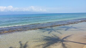 Ein Strand mit klarem Wasser, sandigem Ufer und Palmen im Schatten.