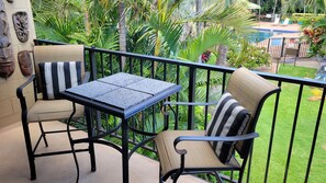 A balcony with two chairs and a table, surrounded by lush greenery and a pool. - Oceanfront Prop Family Friendly Pool 2BD/2BTH AC (Lahaina)