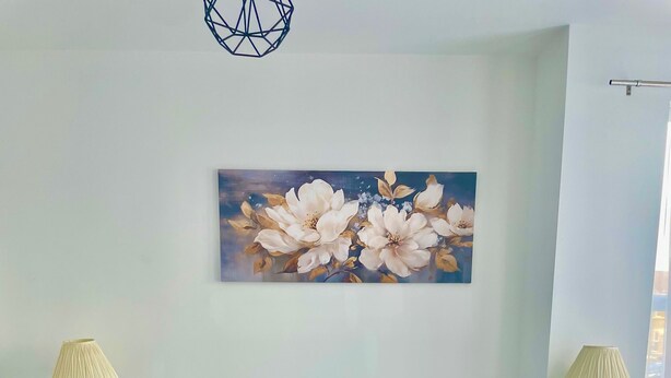 Ein ordentlich bezogenes Bett mit weißen Leinen und einer dunkelblauen Decke, flankiert von zwei Nachttischen mit Lampen, einem an der Wand hängenden Blumenbild und einer Deckenleuchte.