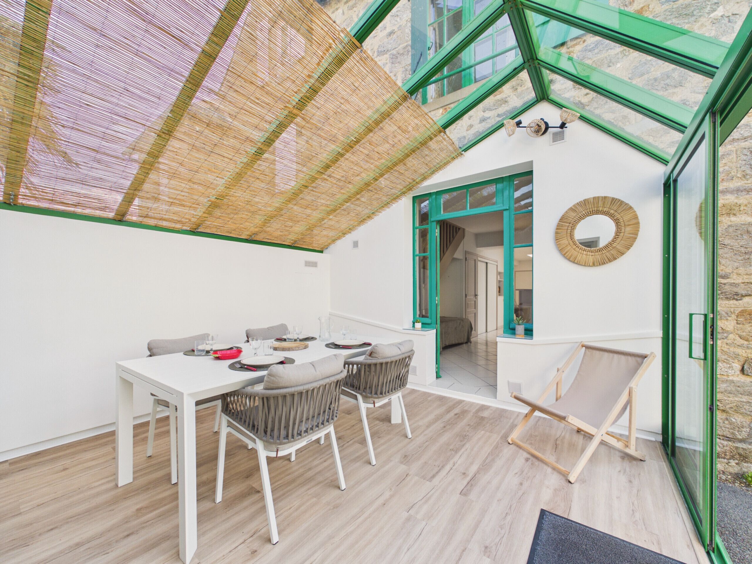 A modern room with a glass ceiling, a dining table with chairs, a wicker chair, a small table with a plant, and a view of the outside.