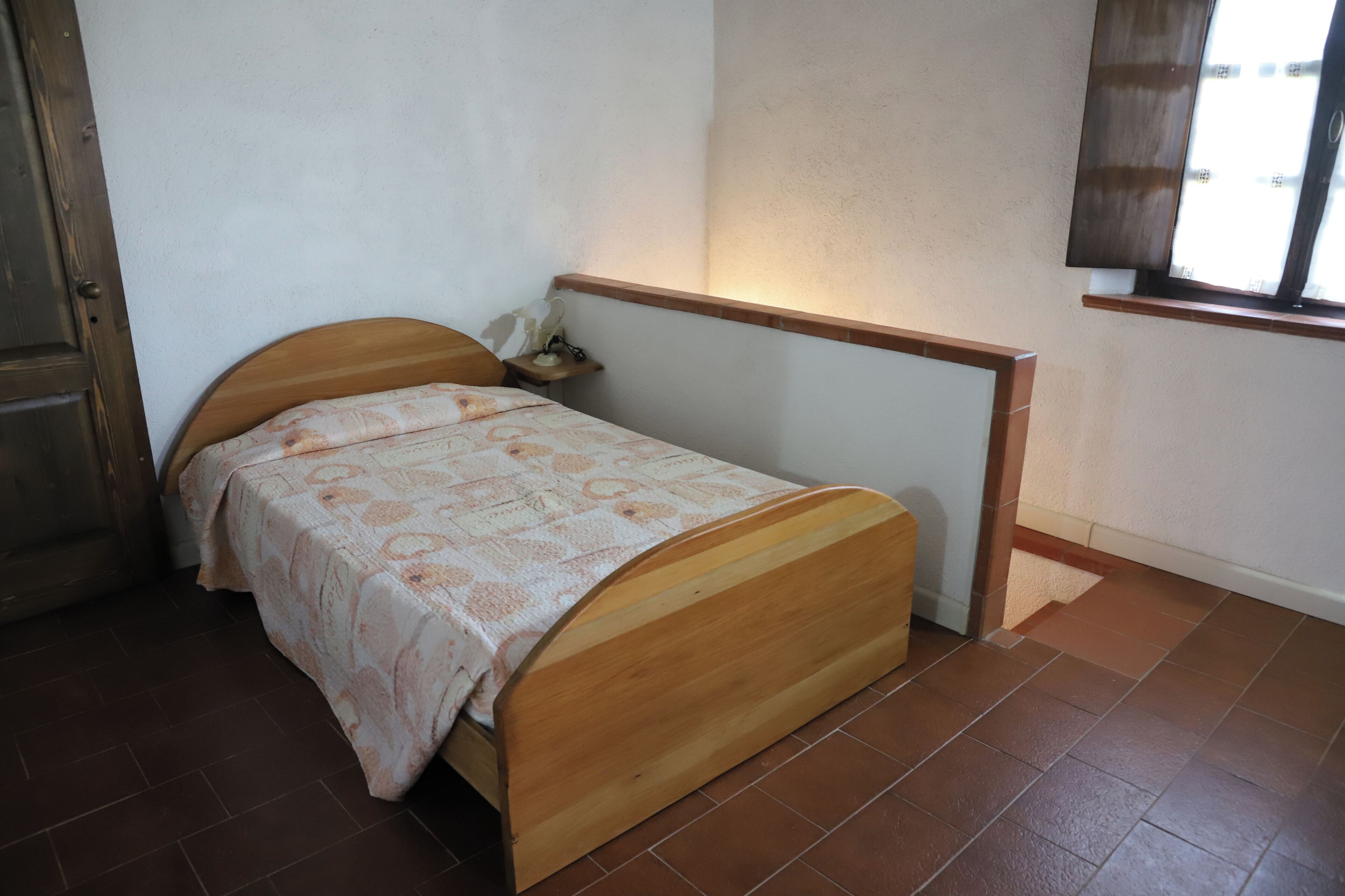 A single bed with a wooden headboard and a patterned bedspread in a room with tiled flooring and a window with curtains.