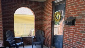 A covered patio area with brick walls, two Adirondack chairs, a small table, and a potted plant with yellow flowers. - Huntington Village House 4BR – Walk to everything (Huntington)
