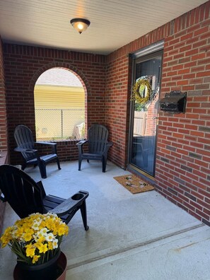 Un patio techado con paredes de ladrillo, dos sillas Adirondack, una mesita y una planta en maceta con flores amarillas.