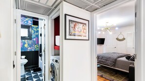 A compact room with a washing machine, a bed with patterned bedding, a framed artwork on the wall, and a ceiling light fixture. - World Cup | .4 Mi to Ponce | 3 Mi to Mercedes Benz (Atlanta)