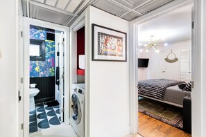 Ein kleines Zimmer mit Waschmaschine, einem Bett mit gemusterten Betttextilien, einem gerahmten Kunstwerk an der Wand und einer Deckenleuchte.