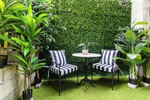 Eine Terrasse mit zwei gestreiften Stühlen, einem kleinen runden Tisch und einer dichten, grünen Hecke im Hintergrund.