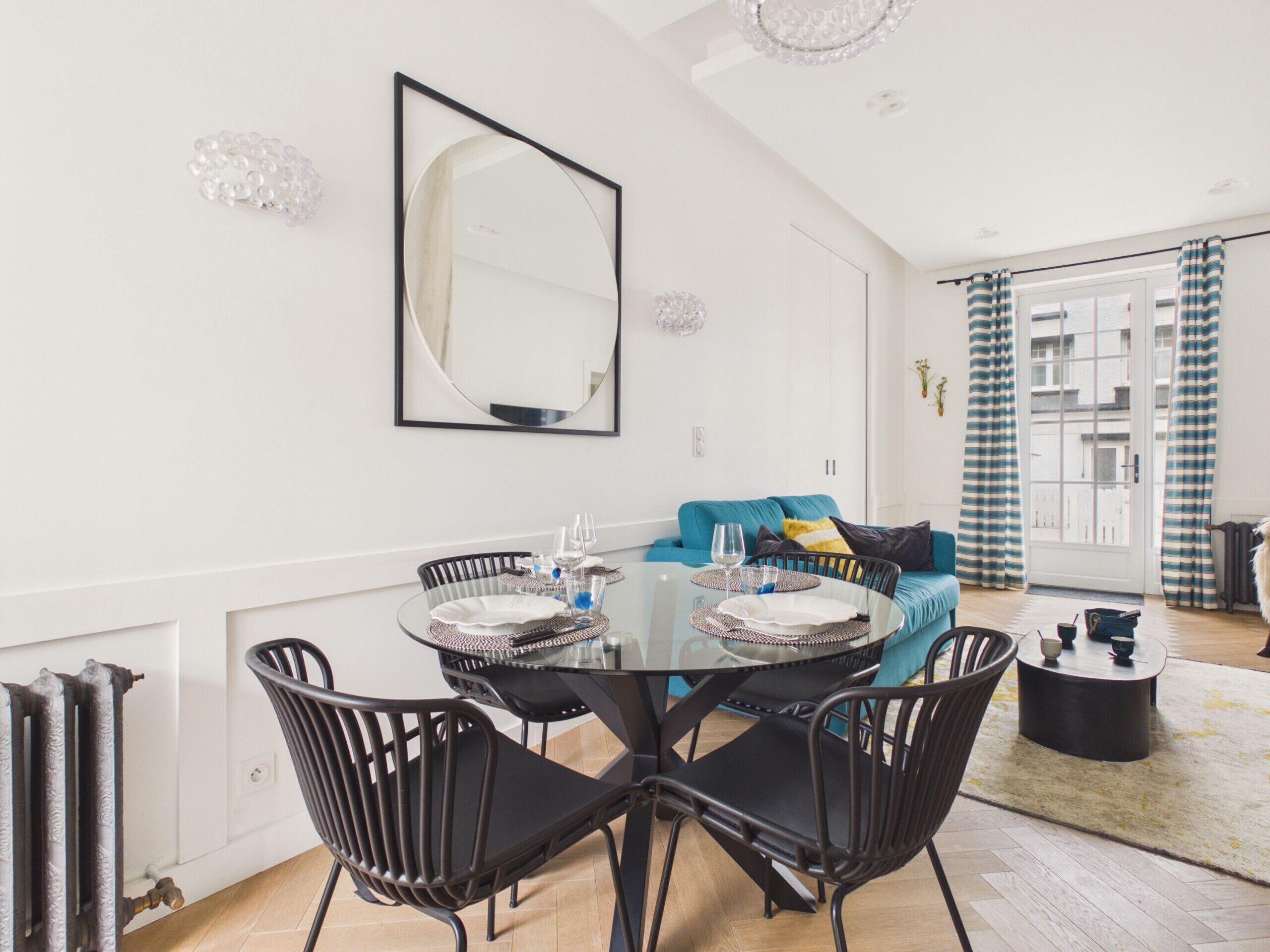 Un comedor con una mesa redonda de cristal, sillas negras, un sofá azul y un espejo grande en la pared.