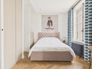 Ein Schlafzimmer mit Bett, Heizkörper, einem Fenster mit Vorhängen und einem Wandbild.