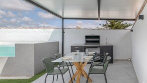 Una zona d'esmorzar exterior moderna amb taula de vidre, cadires metàl·liques i una barbacoa de disseny.