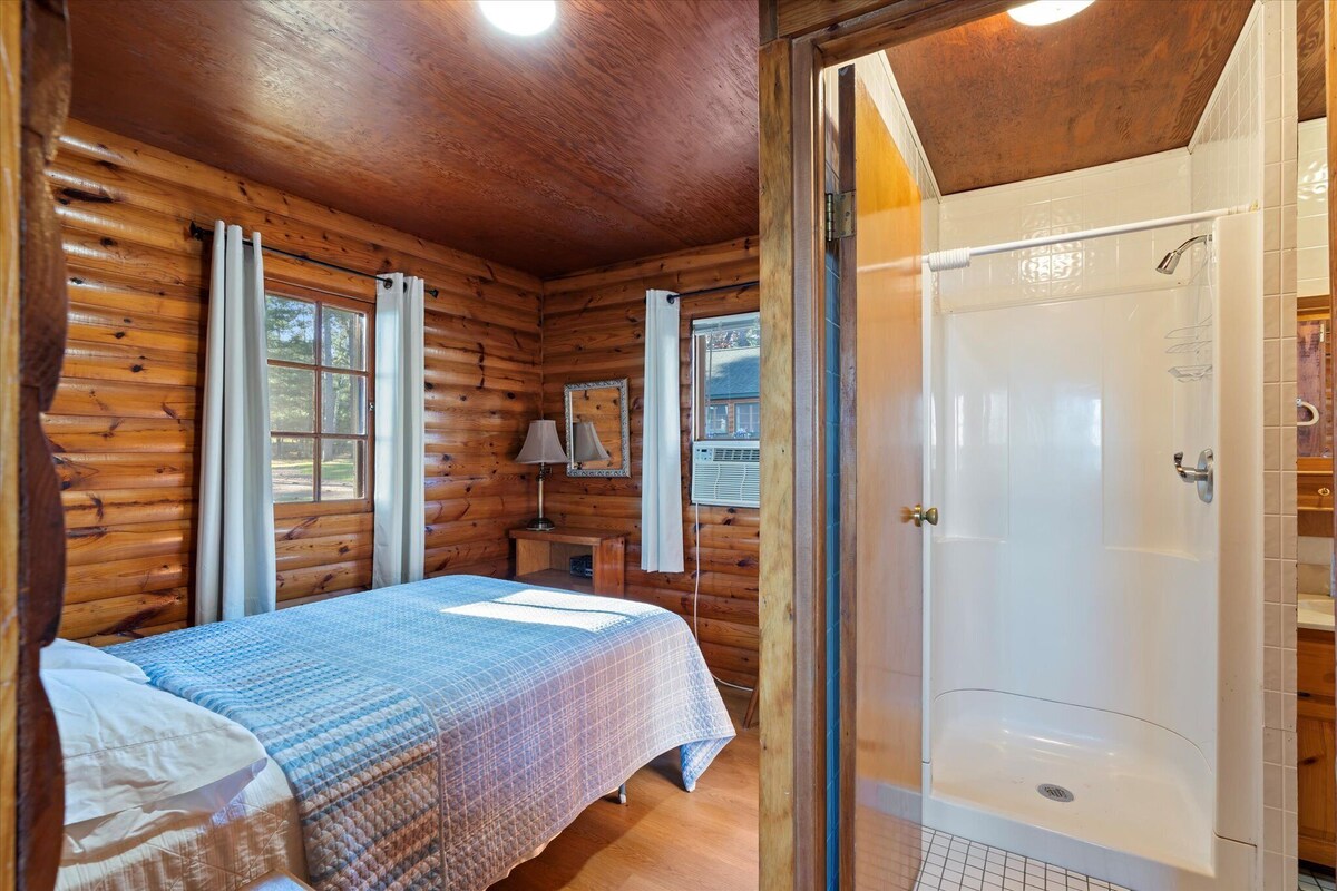 A wooden cabin bedroom with a bed, a window, a nightstand, a mirror, and a shower.