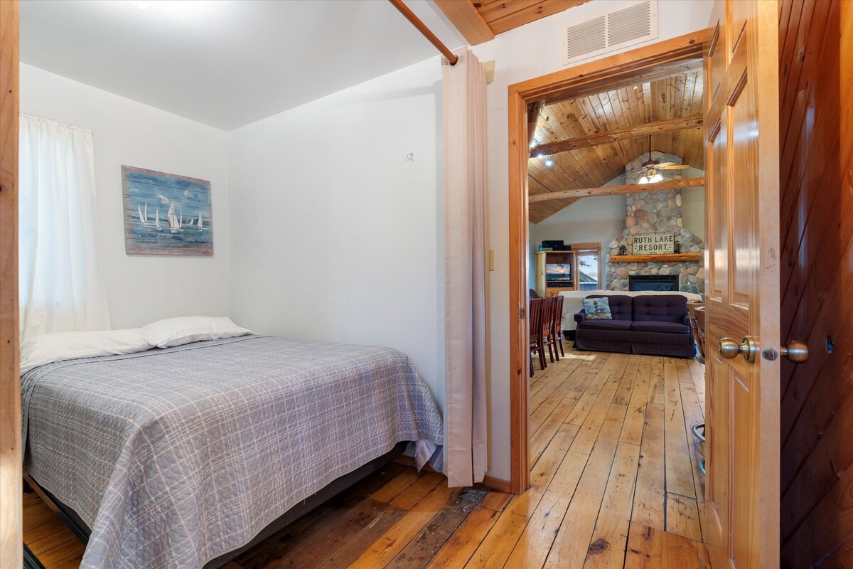 A bedroom with a bed, a painting, and a wooden door leading to a living area with a fireplace.