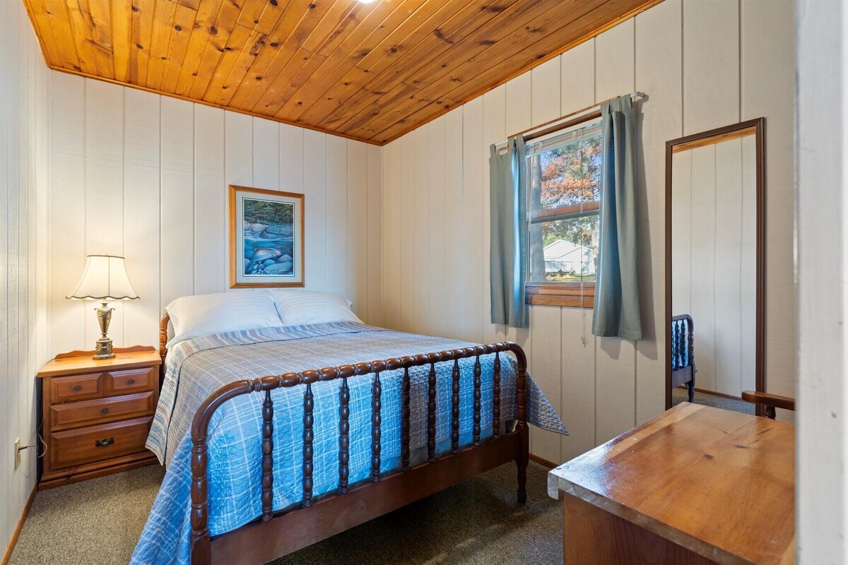 A bedroom with a wooden bed, a nightstand with a lamp, a dresser, a window with curtains, and a mirror.