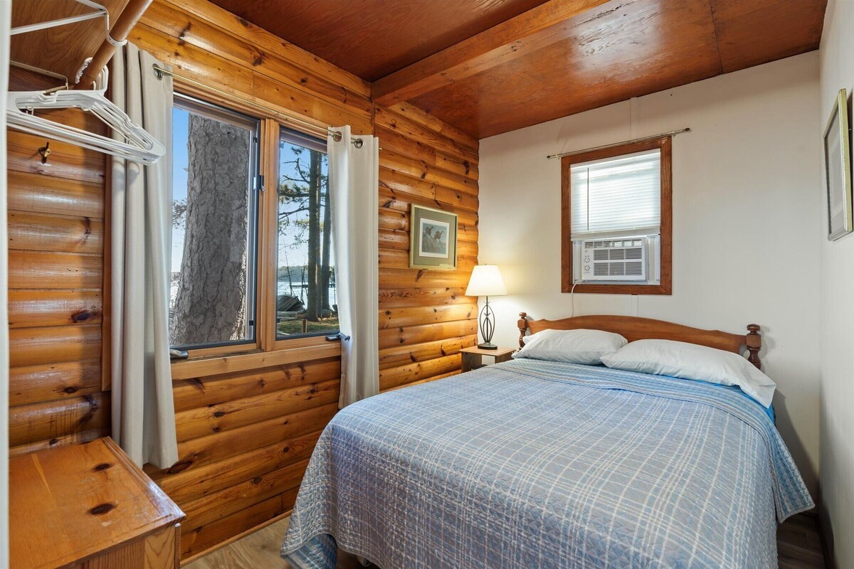 A log cabin bedroom with a bed, a nightstand, a lamp, a window, and a picture on the wall.
