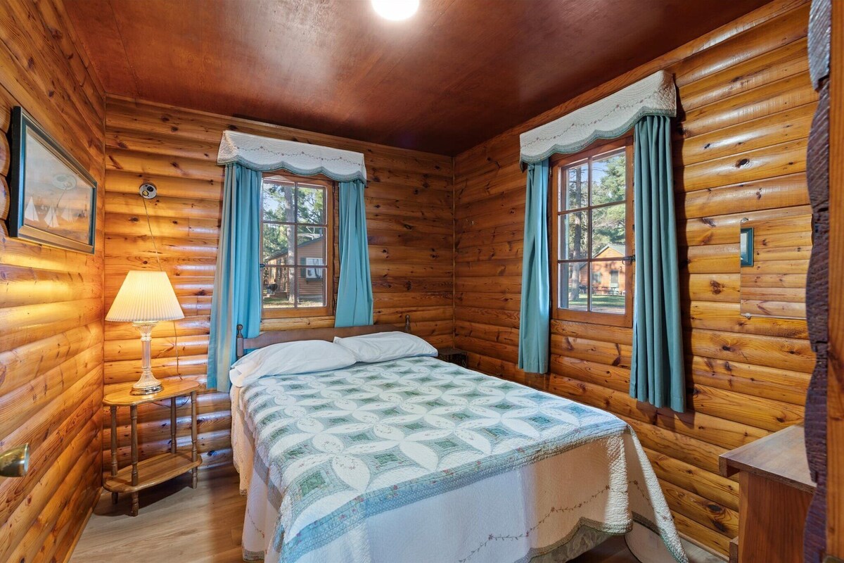 A log cabin room with a bed, two windows with curtains, a lamp, and a small table.