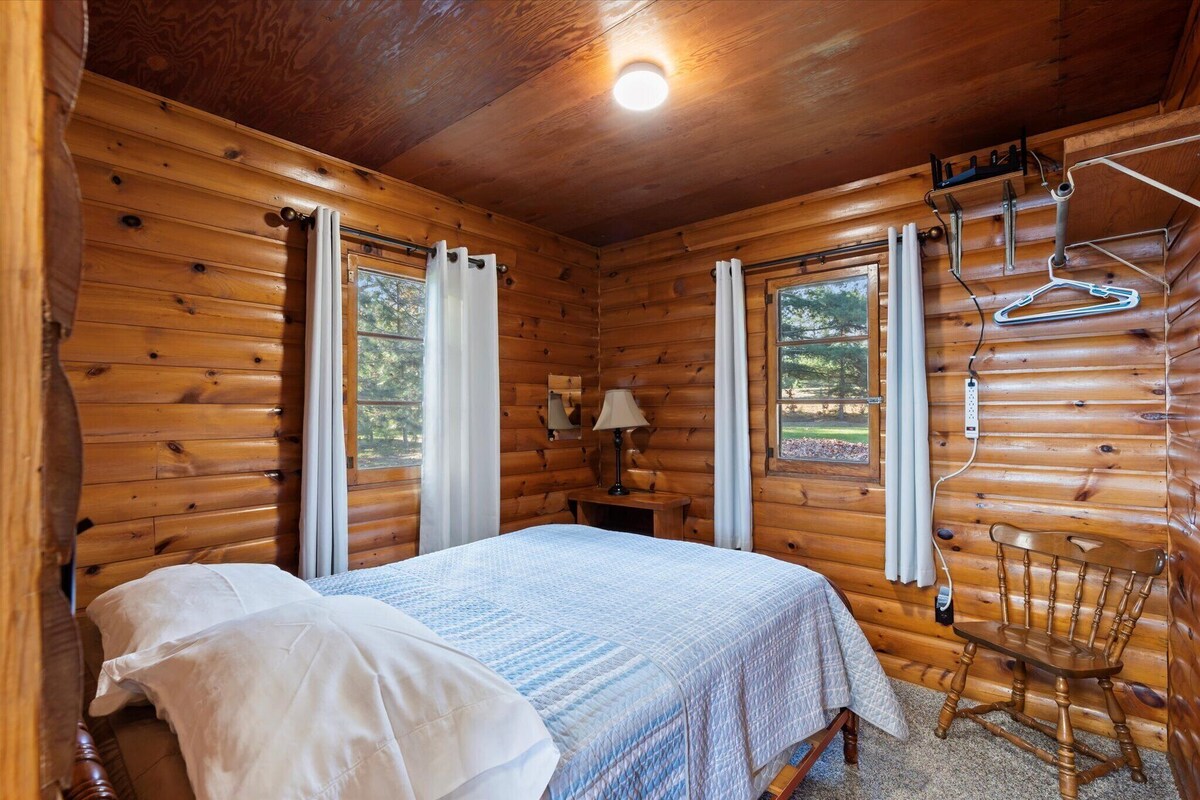 A wooden cabin room with a bed, a chair, a table, and a window.