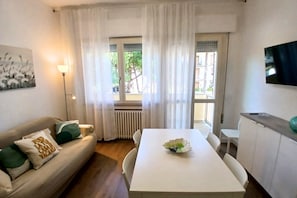 Una habitación con una mesa blanca, un sofá con cojines verdes, una lámpara de pie y un ventanal con cortinas.