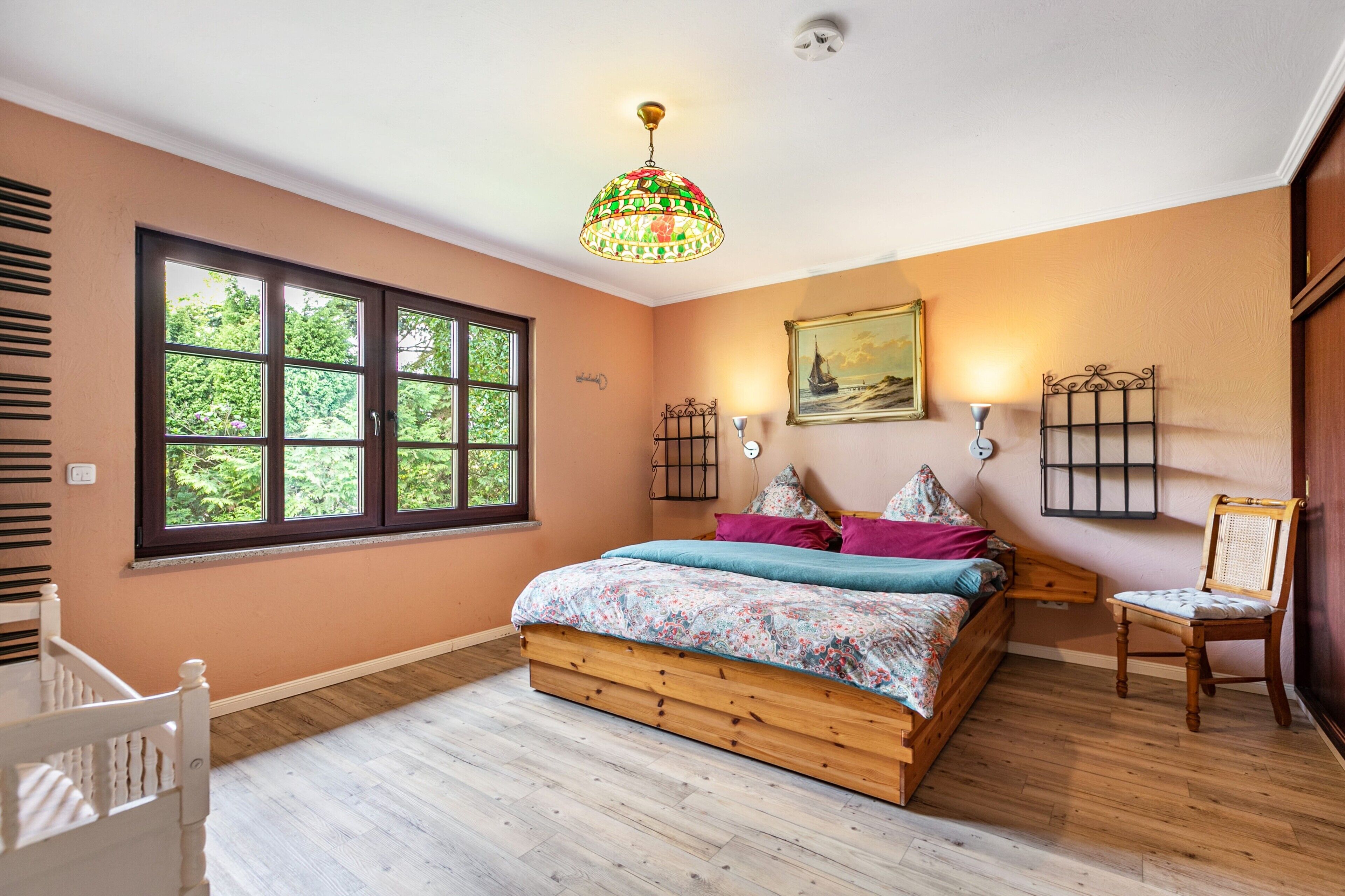 A bedroom with a wooden bed, a window with a view of greenery, a decorative ceiling light, and a wall-mounted picture.