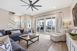 A spacious living room with a sofa, a coffee table, and a white armchair. There is a ceiling fan, a large window with curtains, and a view of the outside. - Just Baskin- Walking Distance-Downtown Morehead City (Morehead City)