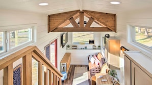 Un loft spacieux avec de hauts plafonds, des poutres en bois, un coin salon confortable avec un canapé et un banc, et de grandes fenêtres offrant une vue sur l’extérieur.