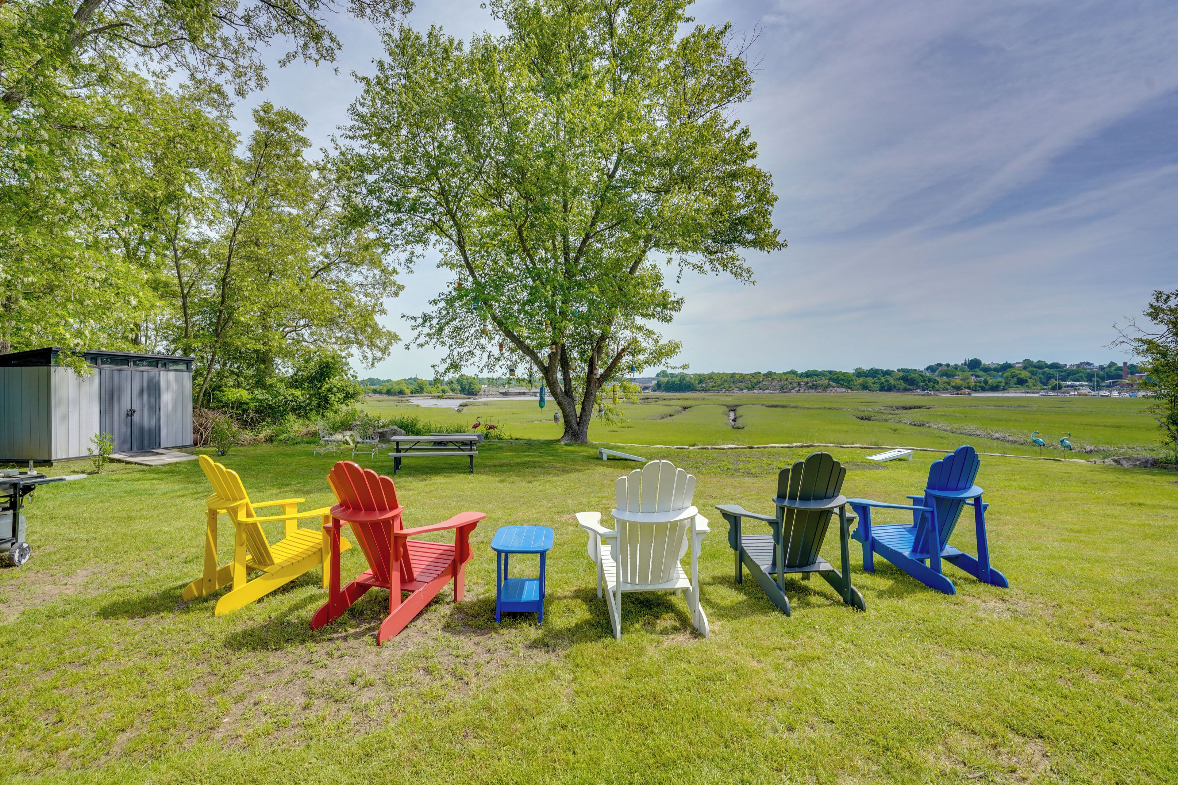Un espace extérieur verdoyant, avec des fauteuils Adirondack de différentes couleurs, une table de pique-nique et un grand arbre.
