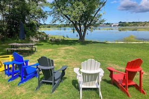 Et græsklædt udendørsareal med Adirondack-stole i blå, grå, hvid og rød, et picnicbord og et træ med fuglehanter.