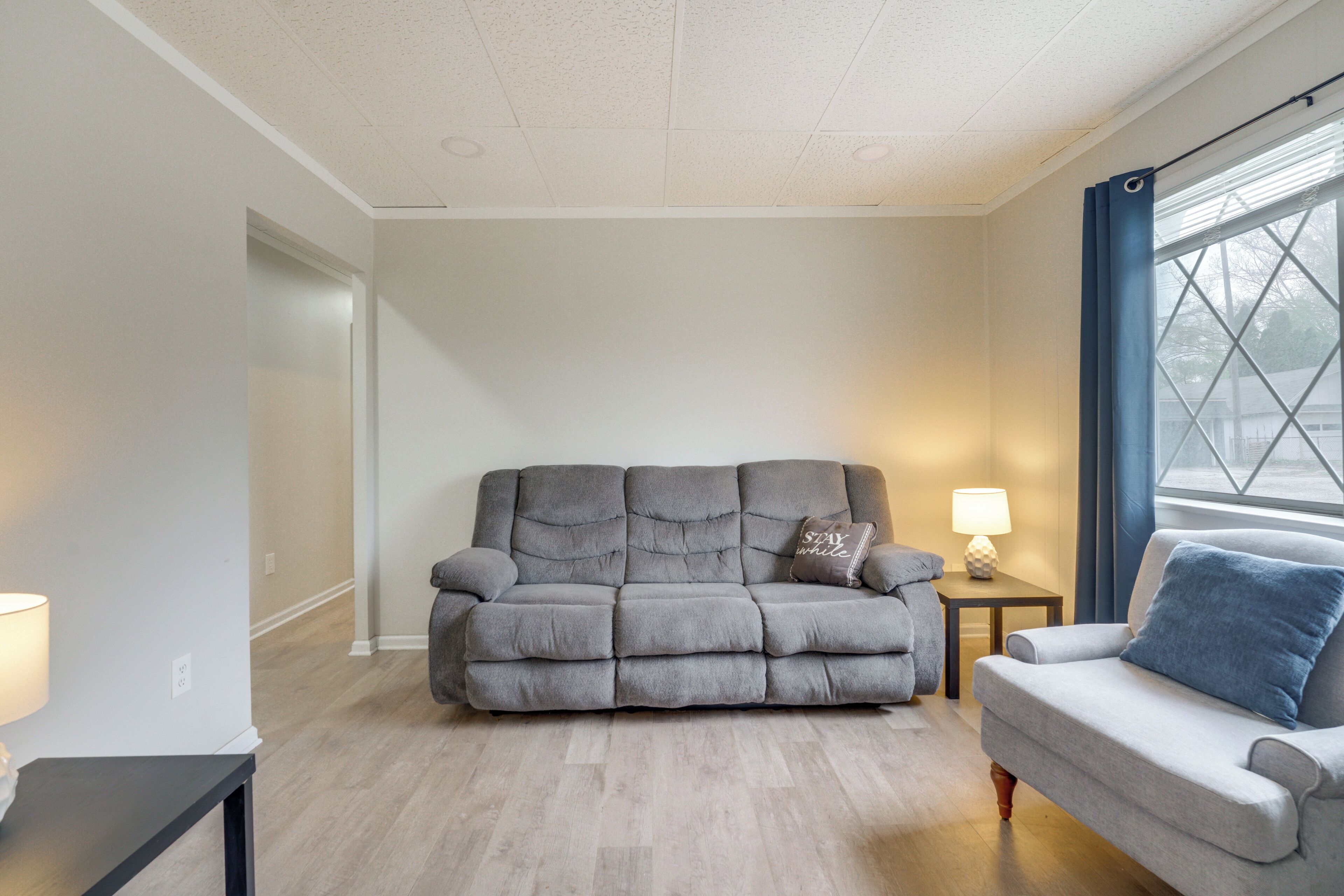A living room with a grey sofa, a smaller grey armchair, a wooden side table with a lamp, and a window with blue curtains.