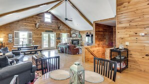 A spacious loft with a high ceiling, wooden walls, a fireplace, a dining area with a table and chairs, and a cozy seating area with a sofa and armchairs. - Game Room, Dock & More! Panoramic Lakefront Oasis (Six Mile)