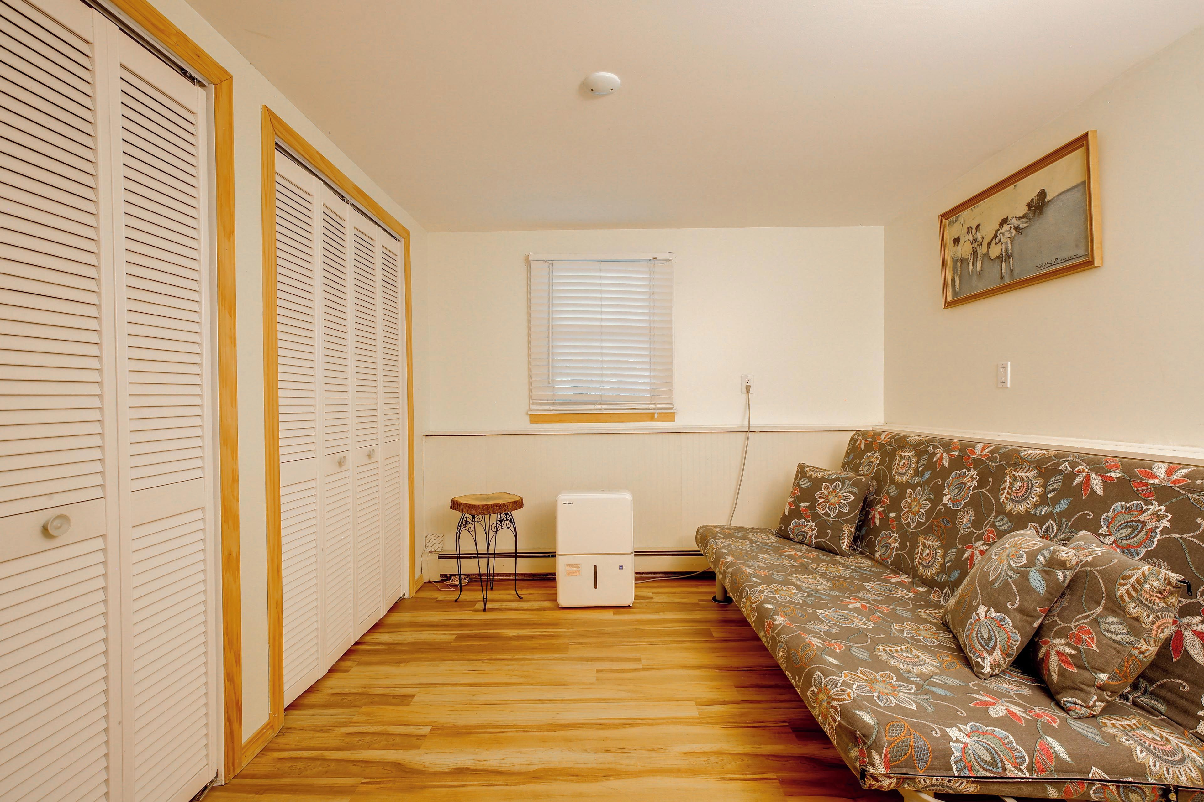 Un espacio habitable compacto con piso de madera, un sofá con tapicería floral, una mesita, un refrigerador blanco y una ventana con persianas.