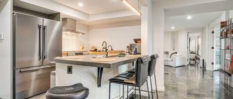 Une cuisine moderne dotée d’un réfrigérateur en acier inoxydable, d’un îlot central avec des tabourets de bar et d’un design épuré et minimaliste.