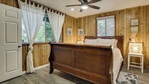 Un dormitorio con pared de paneles de madera, cama, ventilador de techo y ventana con cortinas.