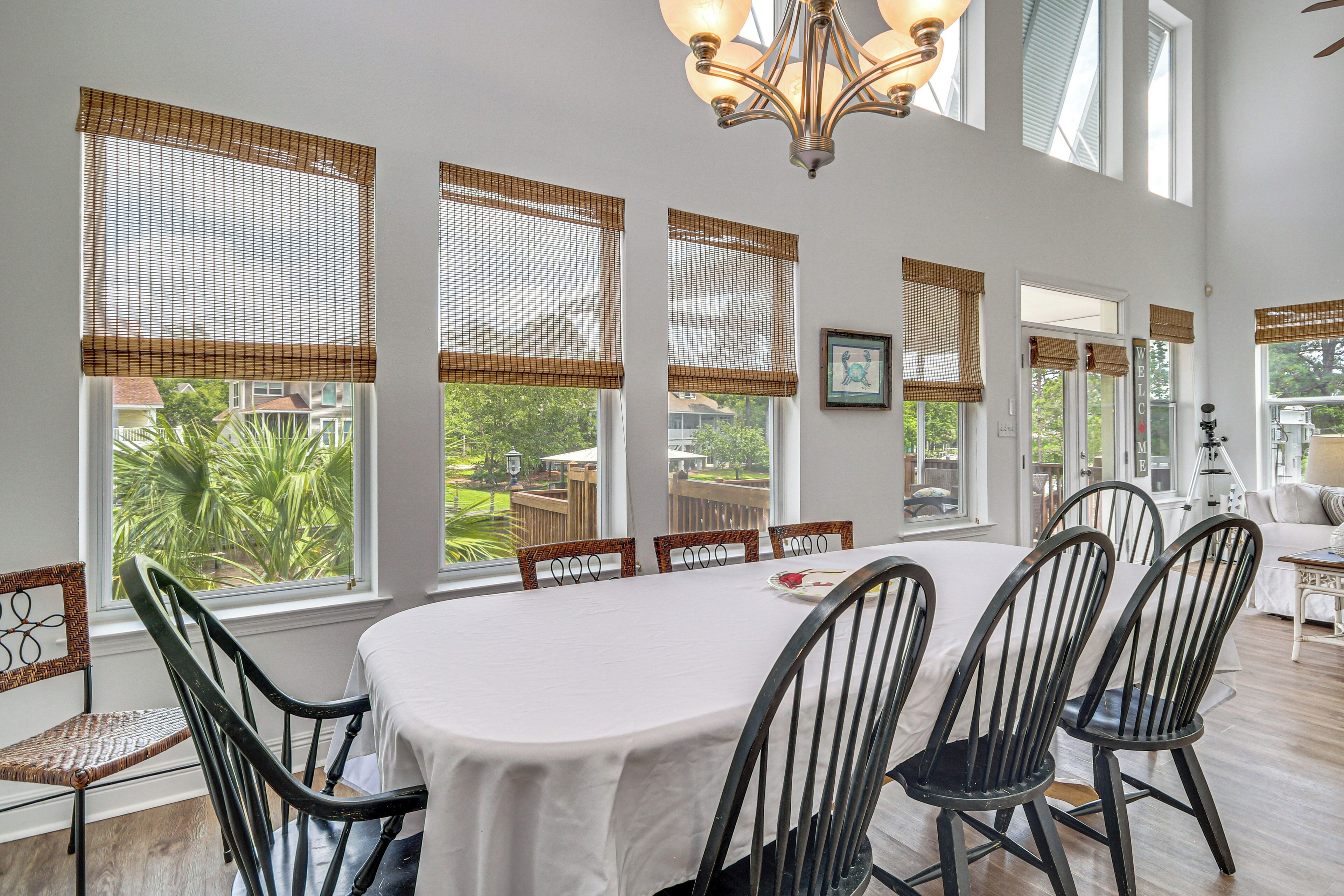 Un coin repas avec une table dressée pour six personnes, entourée de chaises, de grandes fenêtres équipées de stores et d’un lustre.