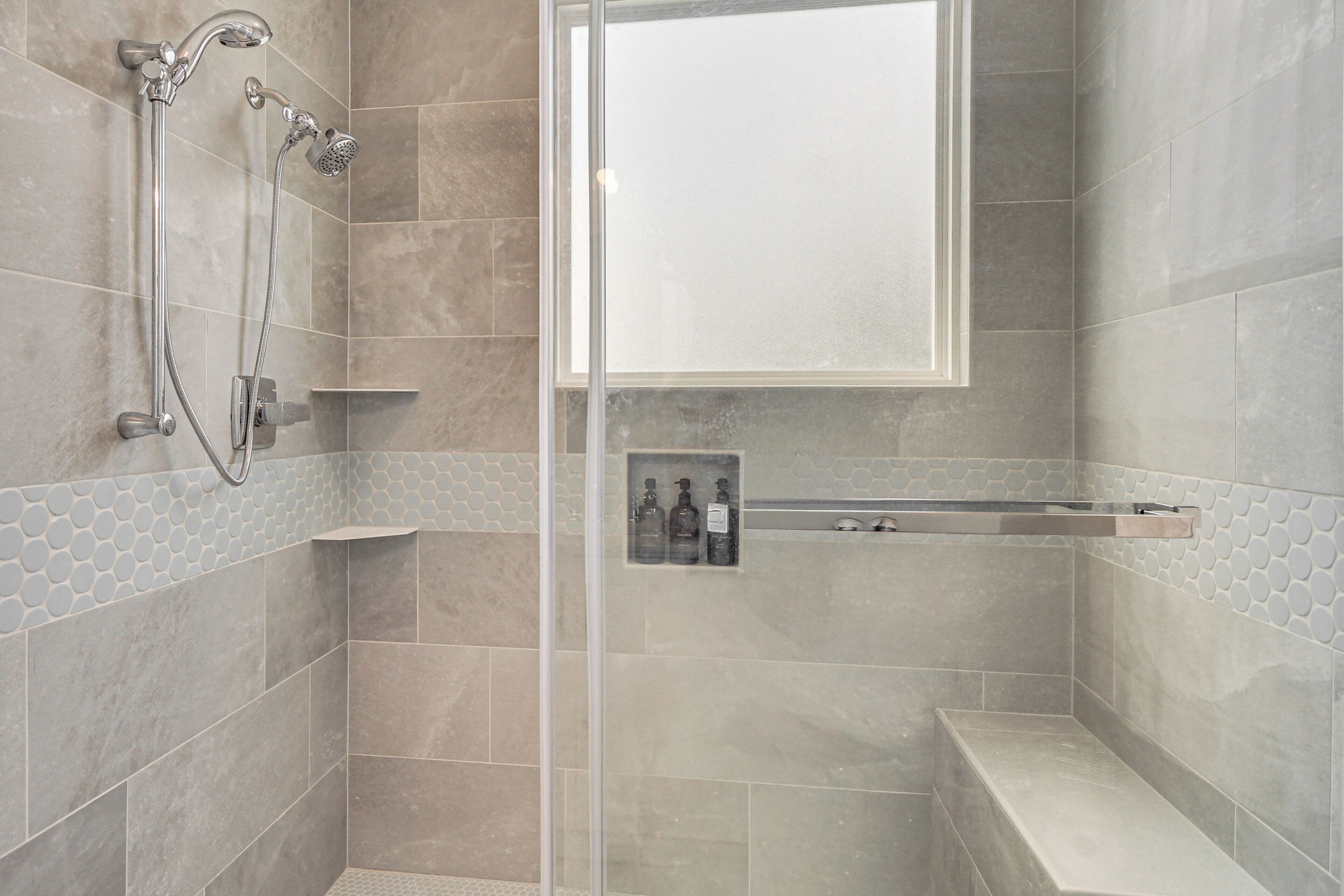 A shower with a glass door, a handheld showerhead, and a wall-mounted showerhead. There is a shelf with bottles and a towel rack.