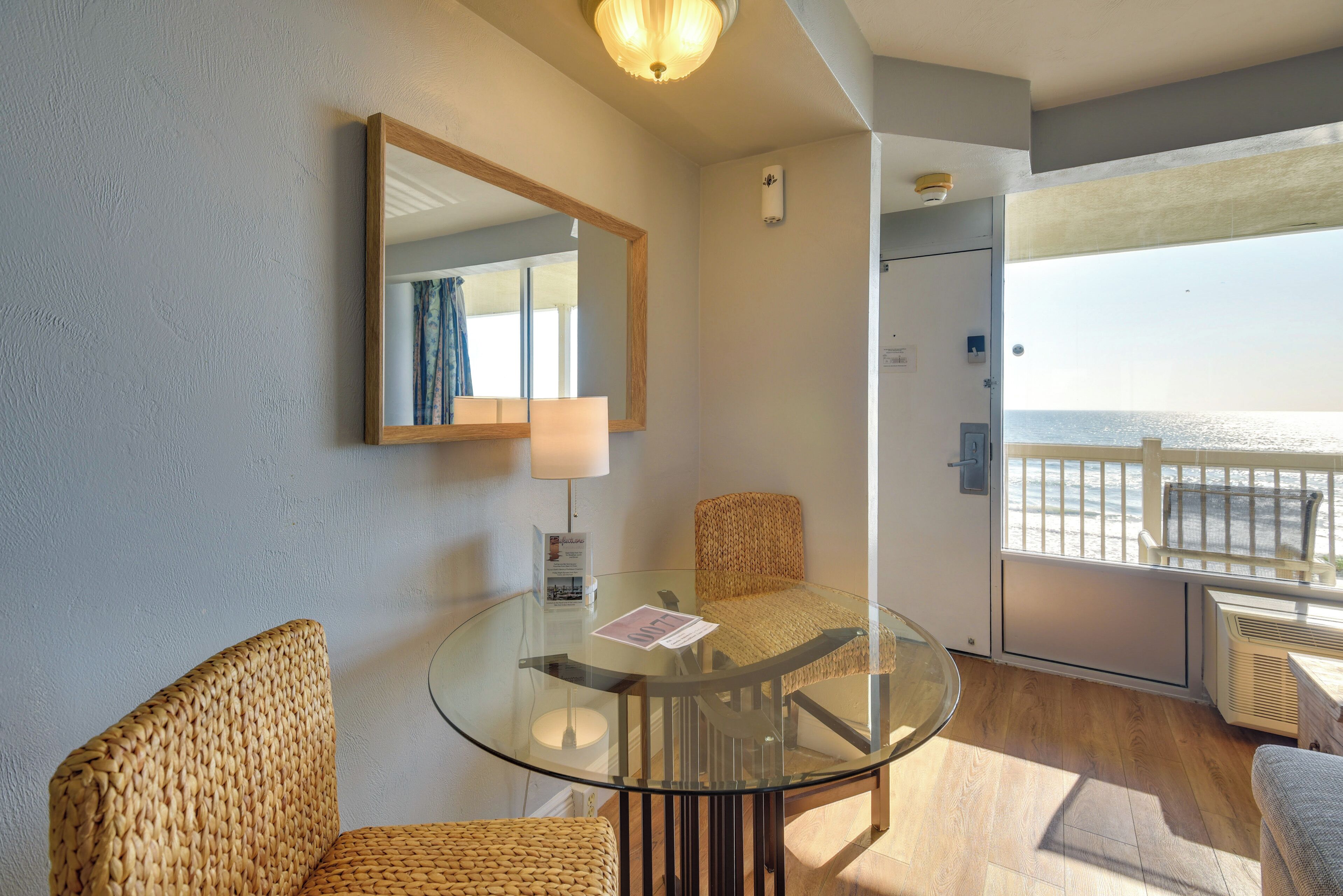 Une pièce avec une table en verre, des chaises en osier, un miroir, une lampe et une vue sur la mer.