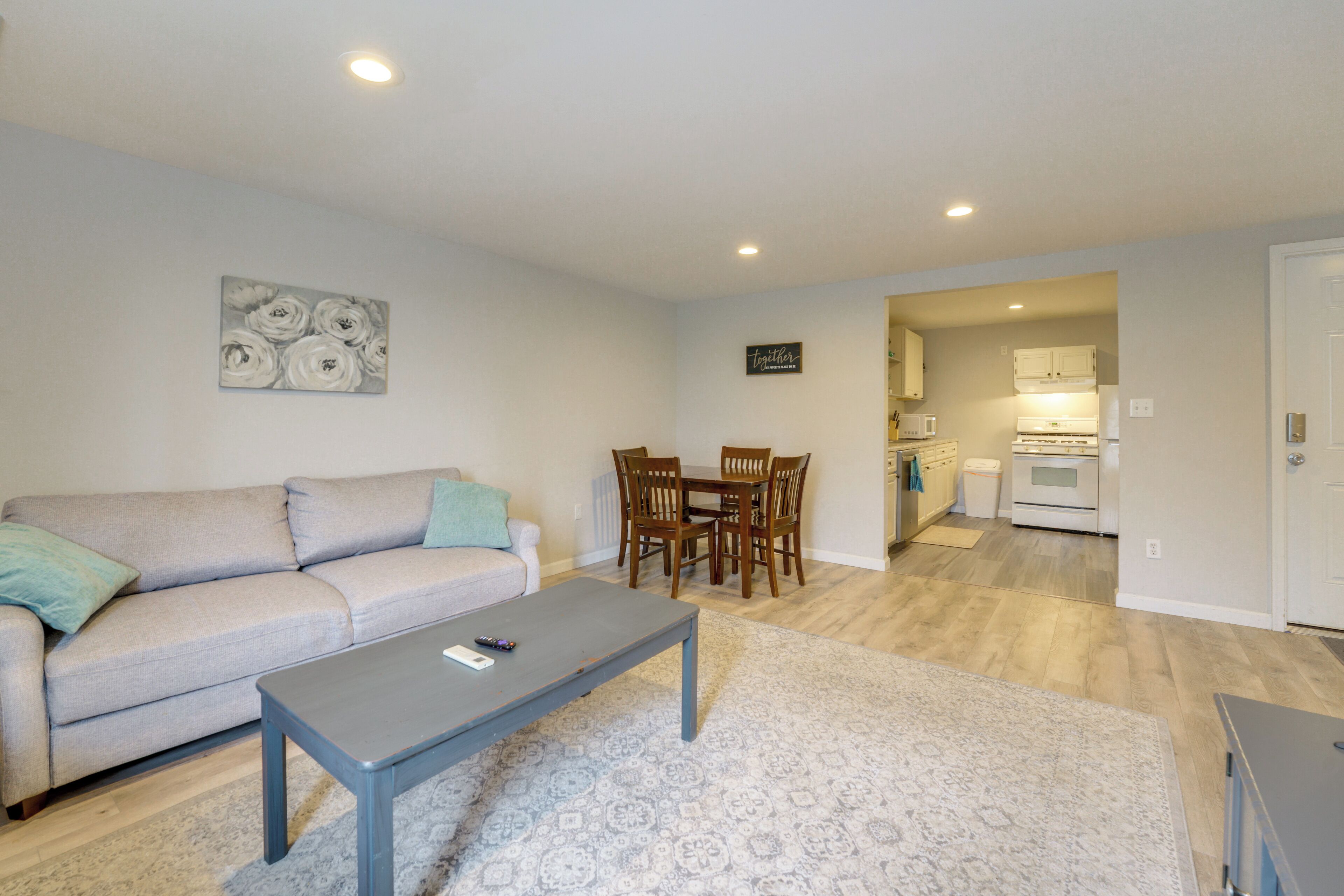 A living room with a sofa, coffee table, and dining table with chairs. There is a kitchen visible in the background.