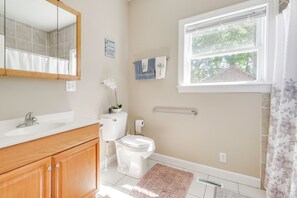 A bathroom with a white sink, wooden cabinets, a toilet, a mirror, and a window with a view of trees. - Soundfront Serenity! Cozy Hertford Home w/ Views (Hertford)