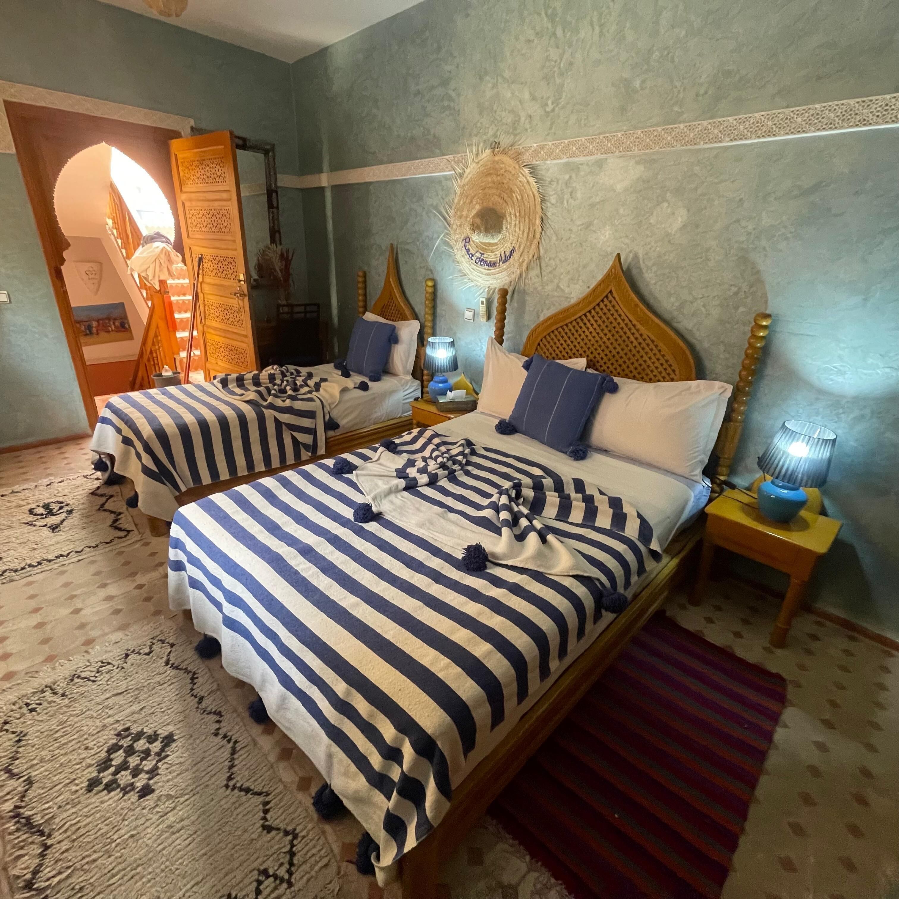 A hotel room with two beds, each with striped bedding and wooden headboards. There is a woven wall hanging above the beds and a small table with a lamp beside one bed.