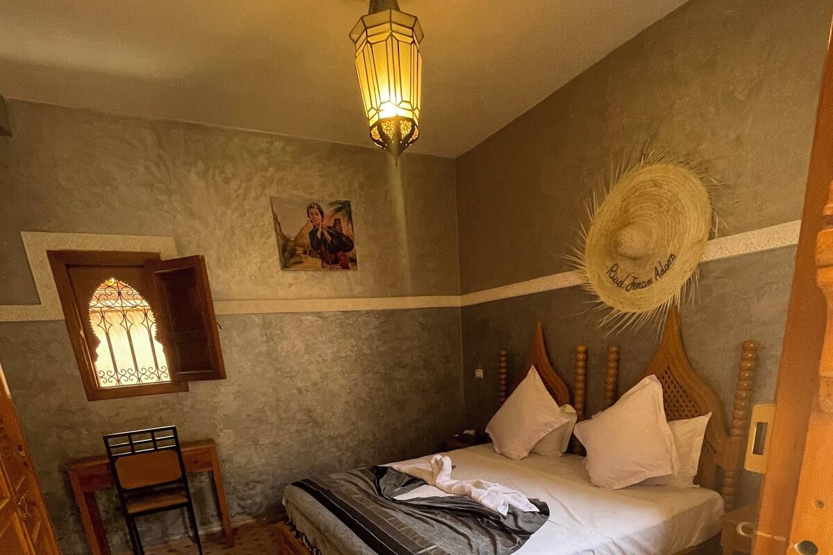 A bedroom with a wooden bed, a small desk, a chair, a decorative wall hanging, and a patterned rug.