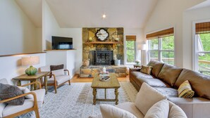 A living room with a stone fireplace, a sofa, a television, and a window with blinds. - Mountain Views & Hot Tub - Your Blue Ridge Getaway (Barnardsville)