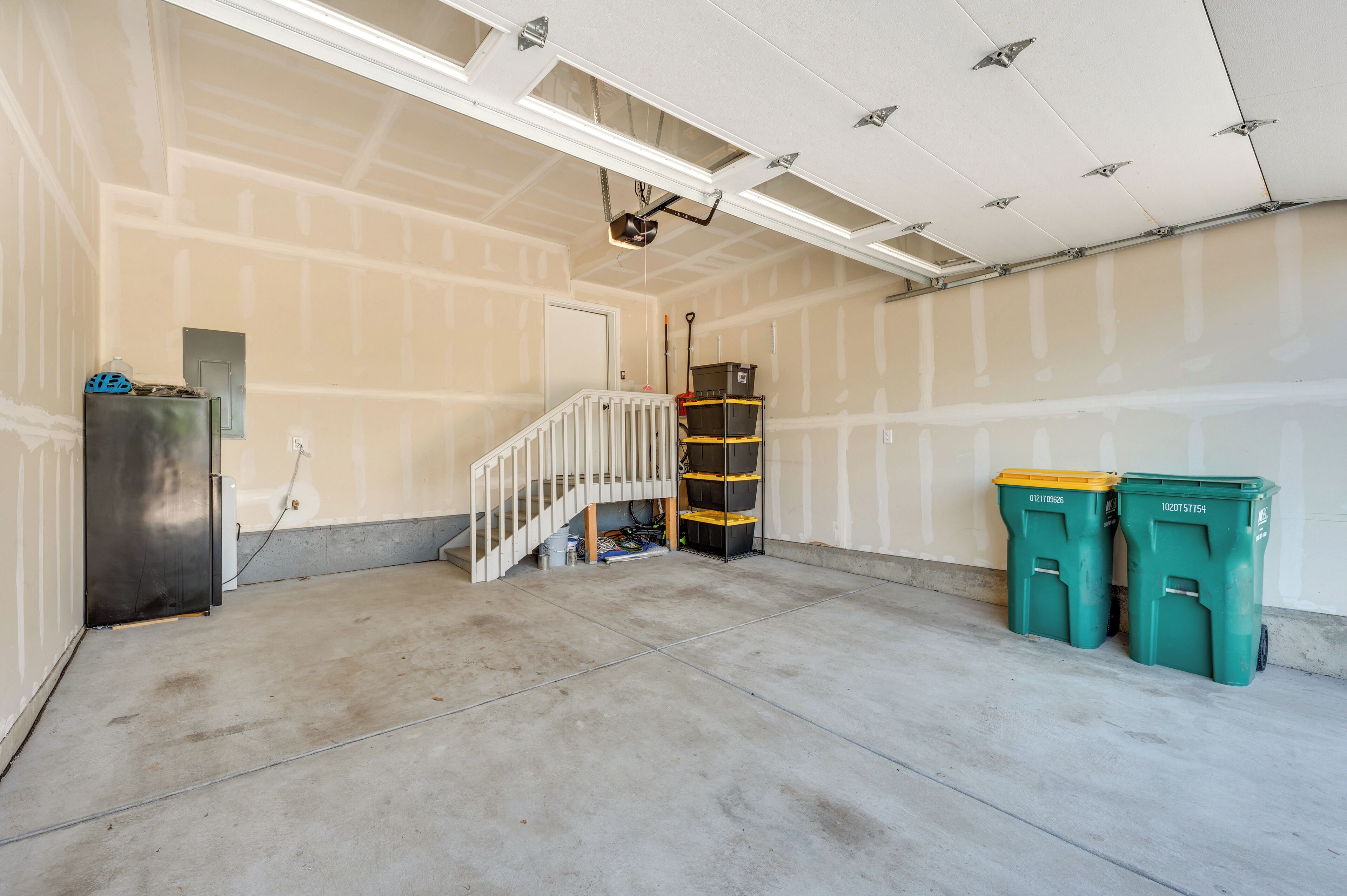 A garage with a concrete floor, a white door, and a white staircase. There are two green trash bins and a black storage unit.