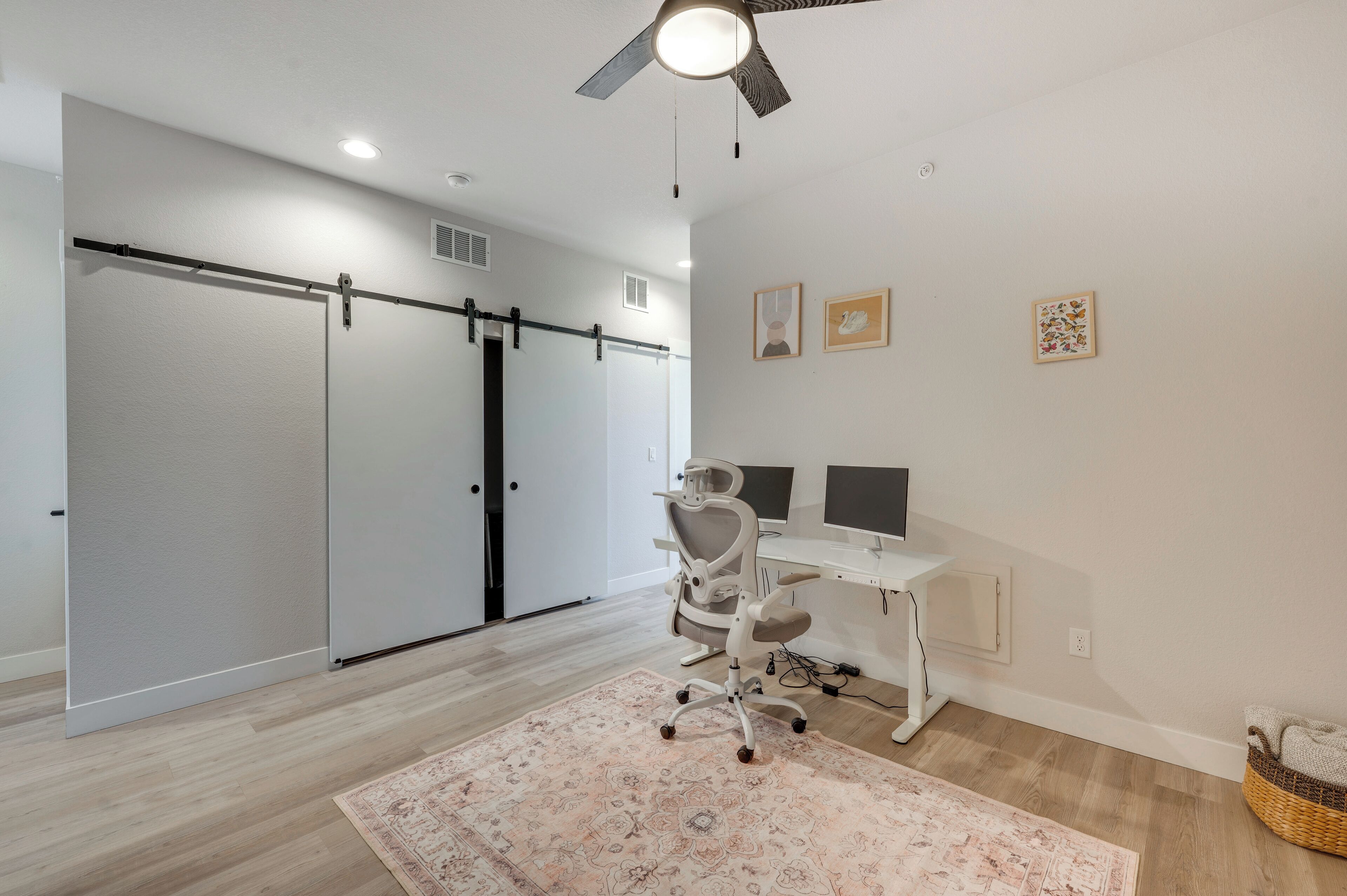 A room with a sliding door, a desk with two monitors, a chair, a ceiling fan, and a patterned rug.