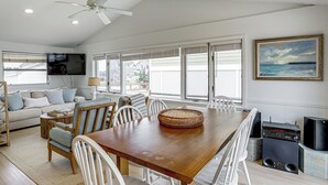 A spacious living area with a wooden dining table, white chairs, a sofa, a television, and a large window with blinds. - Walk to Beach! Pet-friendly Getaway in Lavallette (Lavallette)
