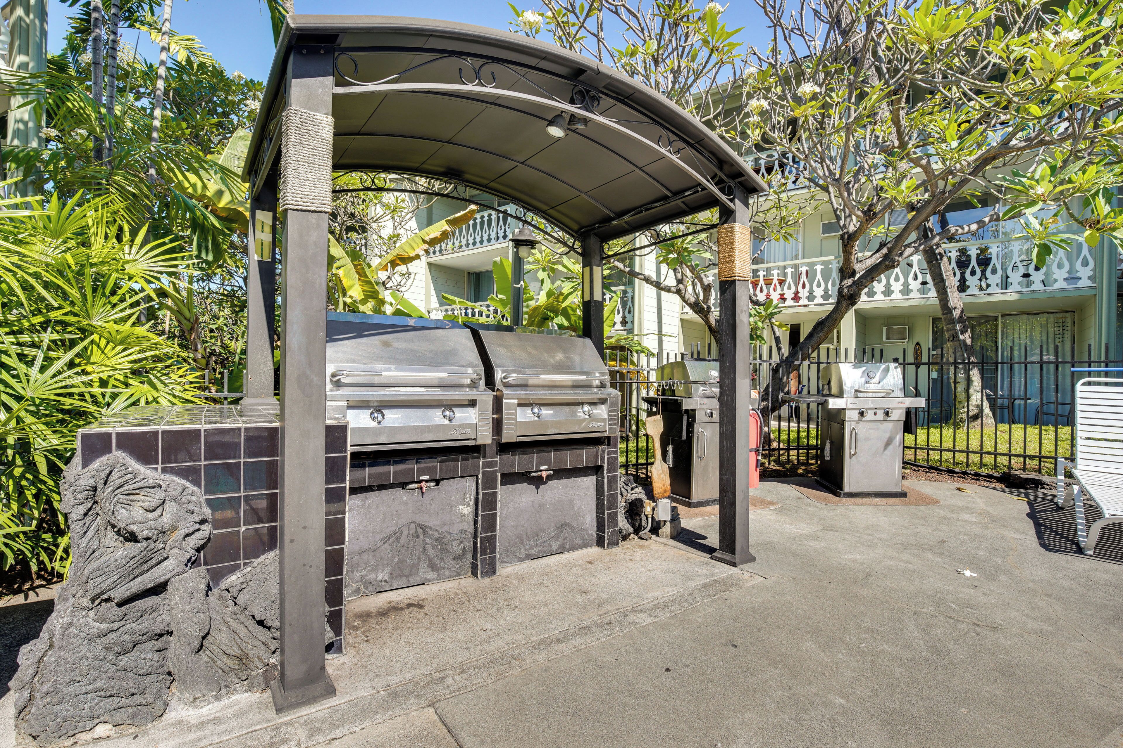 An outdoor grill area with a covered structure, surrounded by greenery and a fenced area.