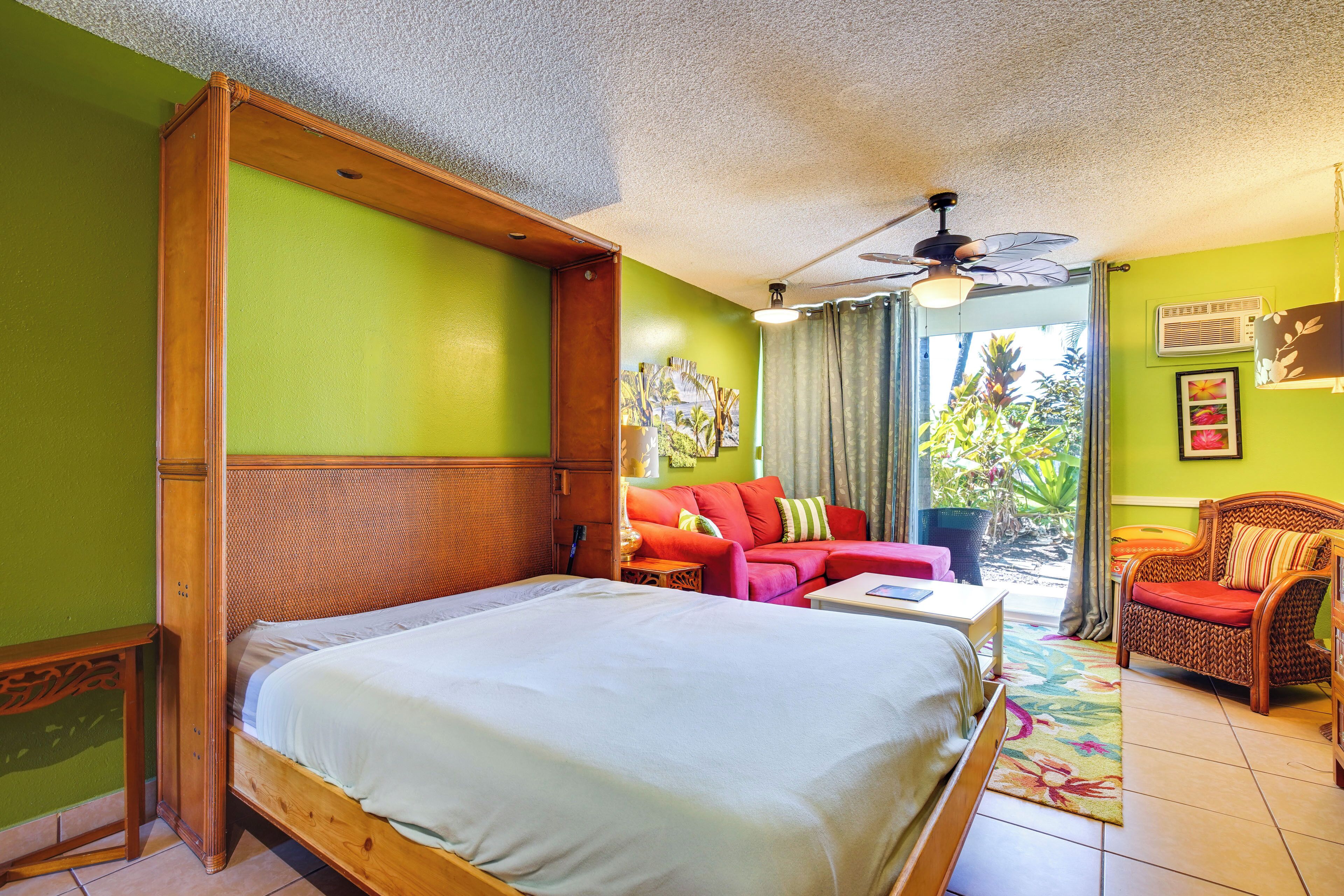 A hotel room with a large bed, a red sofa, a wicker chair, and a ceiling fan.