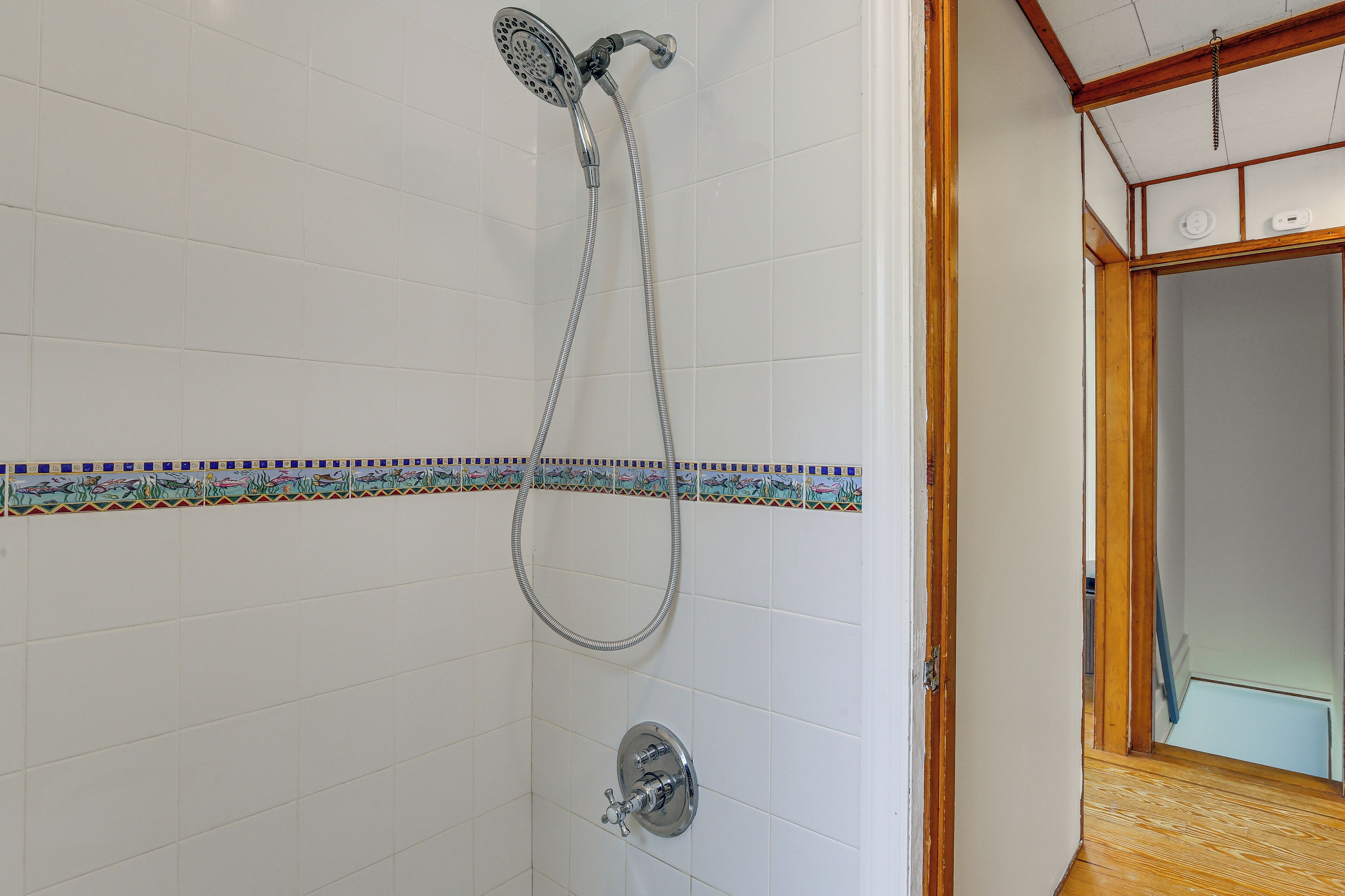 A bathroom with white tiled walls, a shower with a handheld hose, and a decorative tile border.