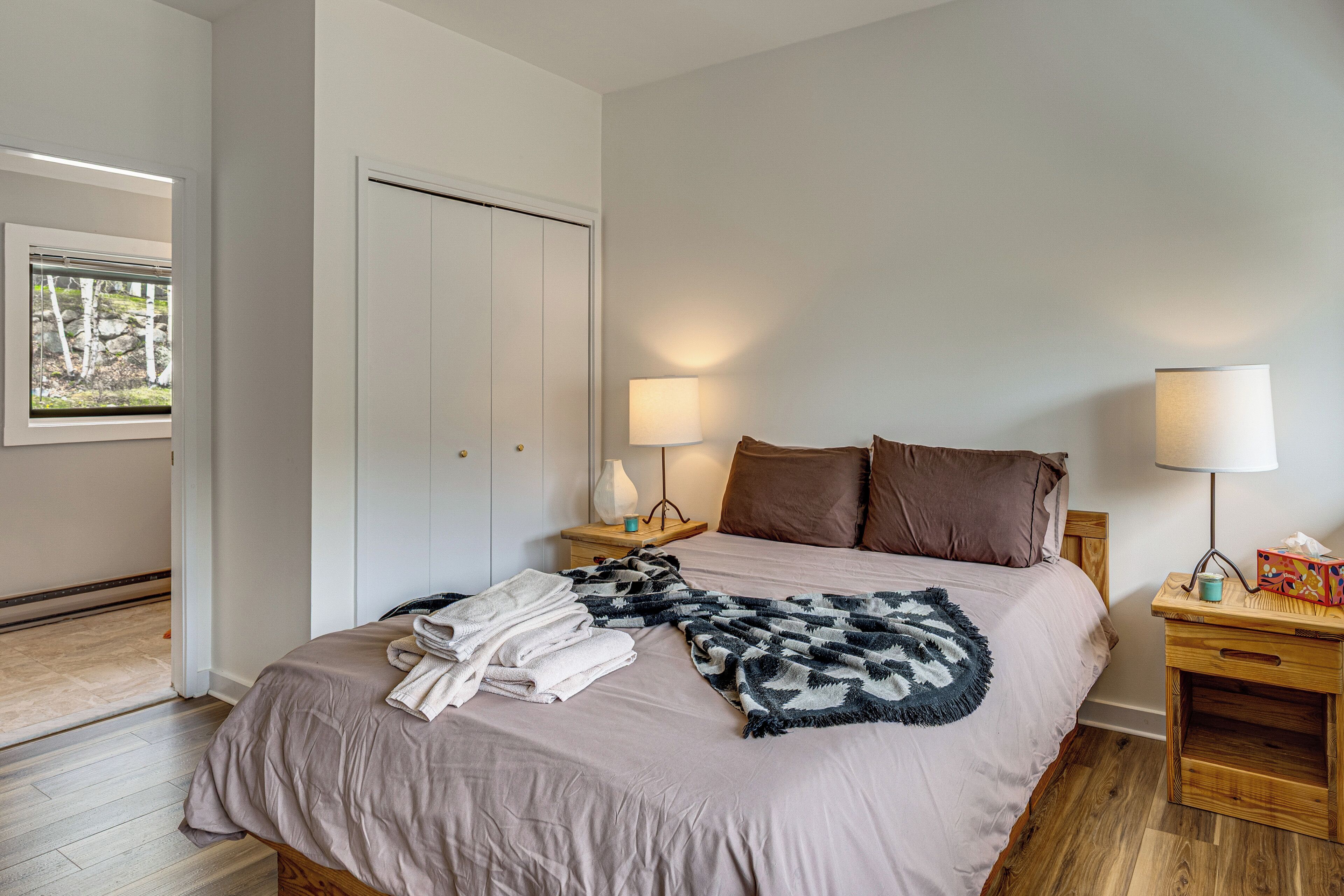 A bedroom with a bed, two bedside tables, a nightstand, a lamp, a window with a view, and a door.