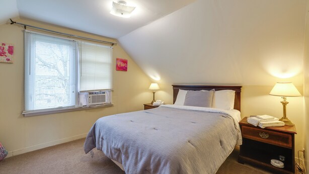 A bedroom with a bed, two bedside tables with lamps, a window with blinds, and an air conditioning unit. - Walk to Falls, Casino! Niagara Family Home w/ Yard (Niagara Falls)