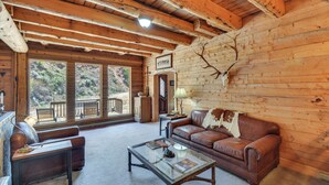 A log cabin interior with a leather sofa, a glass coffee table, and a deer skull mounted on the wall. - Fire Pit: 20-acre Creekfront Retreat in Gunnison! (Gunnison)