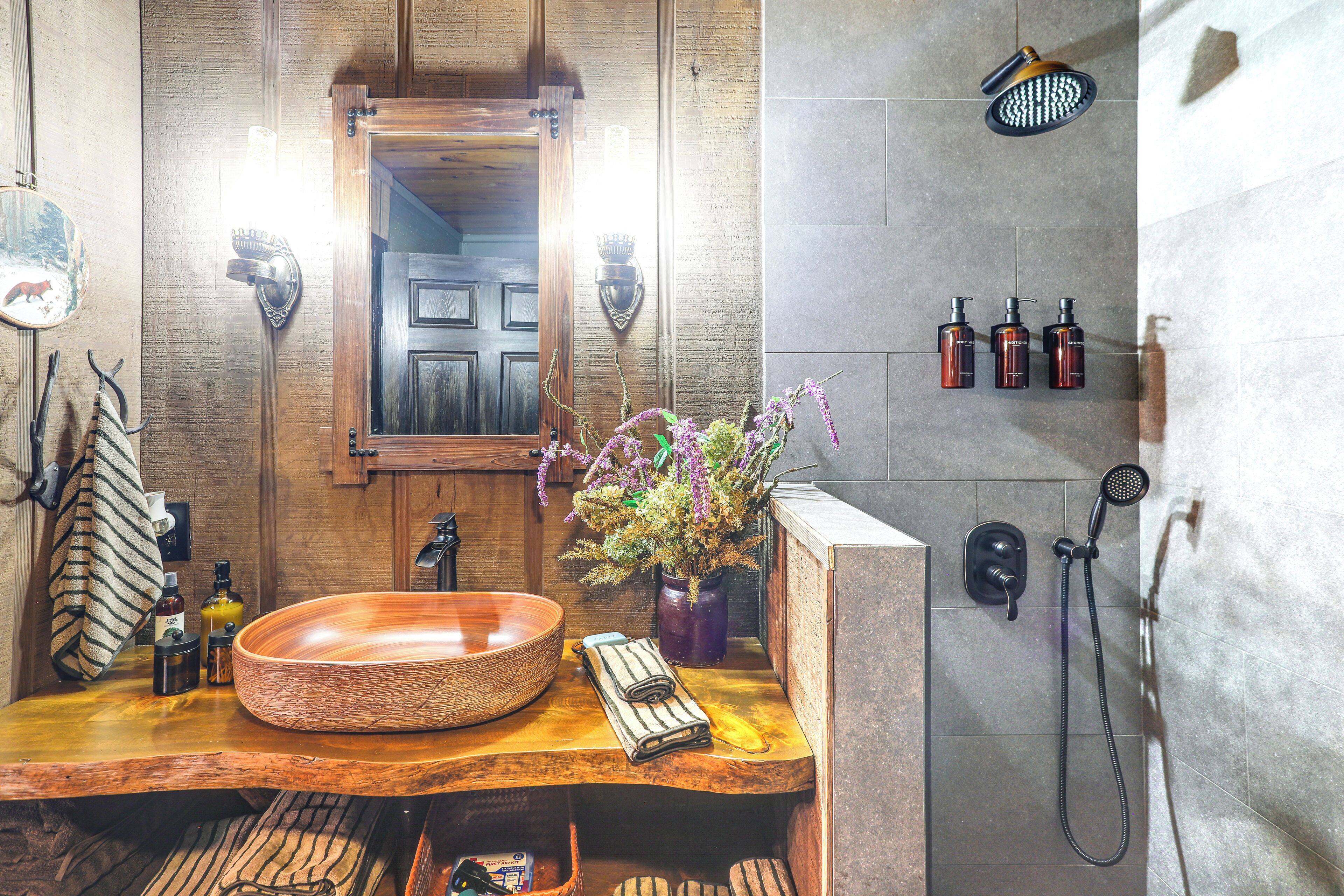 A bathroom with a wooden vanity, a large round sink, a mirror, a shower area with a handheld showerhead, and a wall-mounted towel rack.