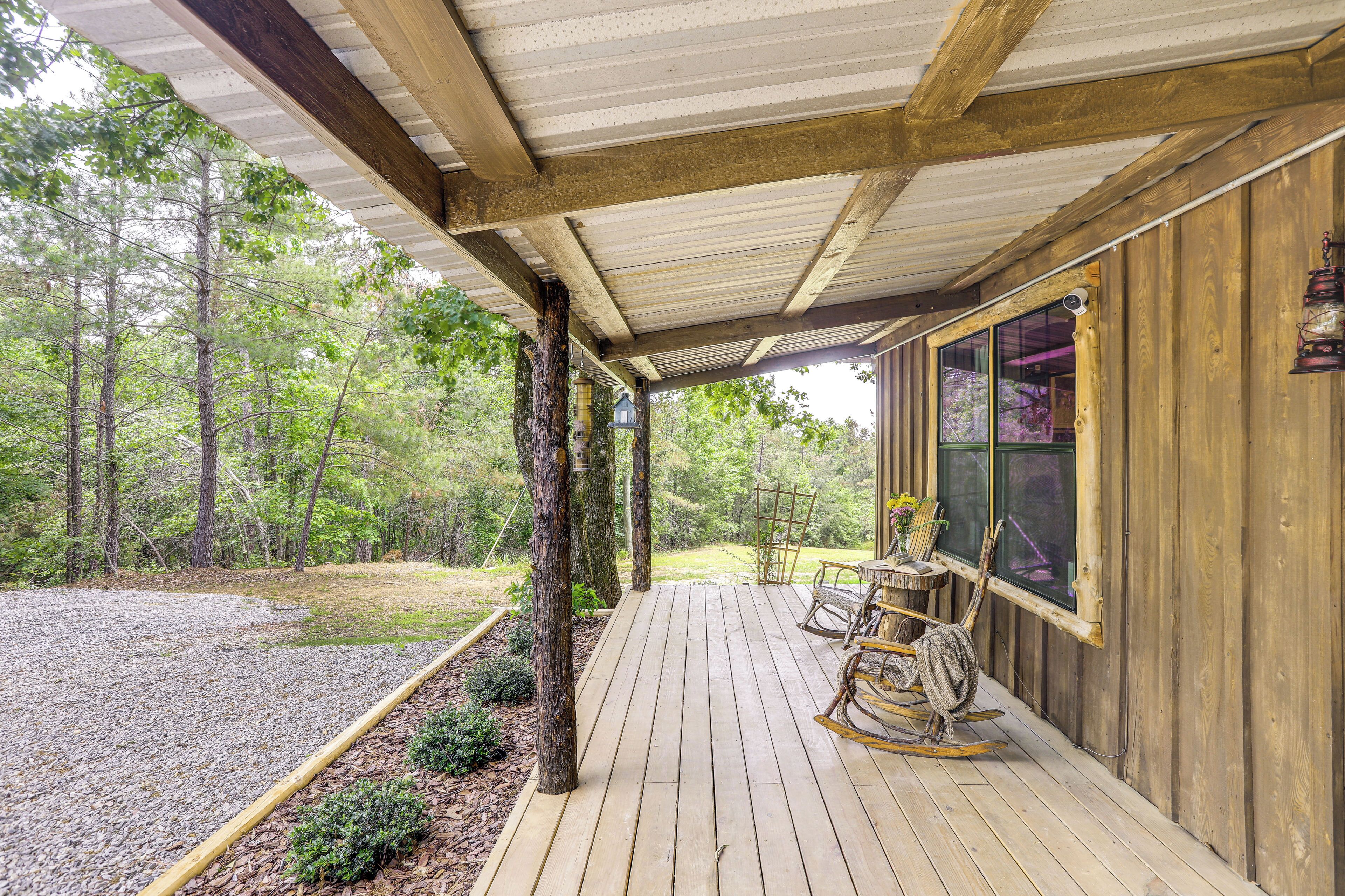 Una terraza de madera con área cubierta, rodeada de árboles, y un sendero de grava que conduce a la entrada.